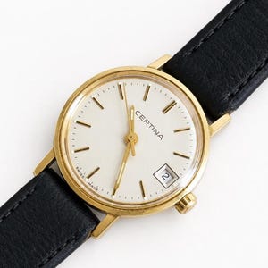 Könnte beinhalten: Eine goldfarbene Certina Armbanduhr mit einem runden weißen Zifferblatt, goldenen Stundenmarkierungen und Zeigern. Die Uhr verfügt über ein Datumsfenster auf der Drei-Uhr-Position und ein schwarzes Lederarmband mit sichtbaren Nähten.