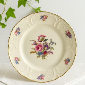 Puede incluir: Plato de porcelana color crema con borde dorado y diseño floral. El plato presenta un ramo central de flores rosas, moradas y azules, con racimos florales más pequeños alrededor del borde. El borde tiene un borde festoneado.