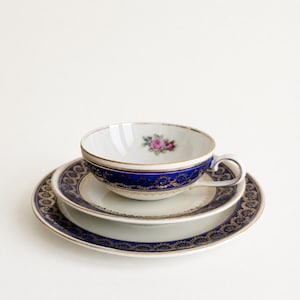 Może przedstawiać: Zestaw białej porcelany składający się z filiżanki i spodka, ozdobiony głębokim niebieskim pasem ze skomplikowanymi złotymi wzorami filigranowymi. Wnętrze filiżanki posiada mały motyw różanej róży, a brzegi wykończone są cienką złotą linią.
