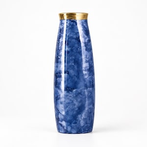 Op de afbeelding: Een hoge, cilindrische vaas met een diepblauw en wit marmerpatroon over het hele lichaam. De bovenrand van de vaas is afgewerkt met een gladde, gepolijste goudkleurige band, wat een duidelijk contrast vormt met het getextureerde blauwe oppervlak.