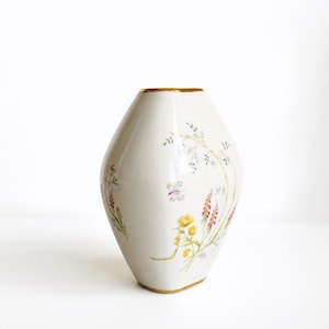 May include: A white porcelain vase with a tapered, angular shape and a gold-trimmed rim and base. The surface is decorated with delicate, hand-painted botanical illustrations featuring yellow flowers, pink foxgloves, and a small butterfly.