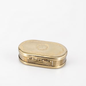 Puede incluir: Una caja de metal de tono dorado con forma ovalada y tapa con bisagras. La superficie superior presenta un borde floral grabado y un motivo circular central. La caja tiene un acabado pulido y un mecanismo de bisagra visible en el borde.