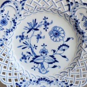 May include: A white porcelain plate featuring a blue floral pattern in the centre and a decorative lattice-style openwork border. The design includes intricate blue painted flowers, leaves, and stems, with scalloped edges and blue accents.