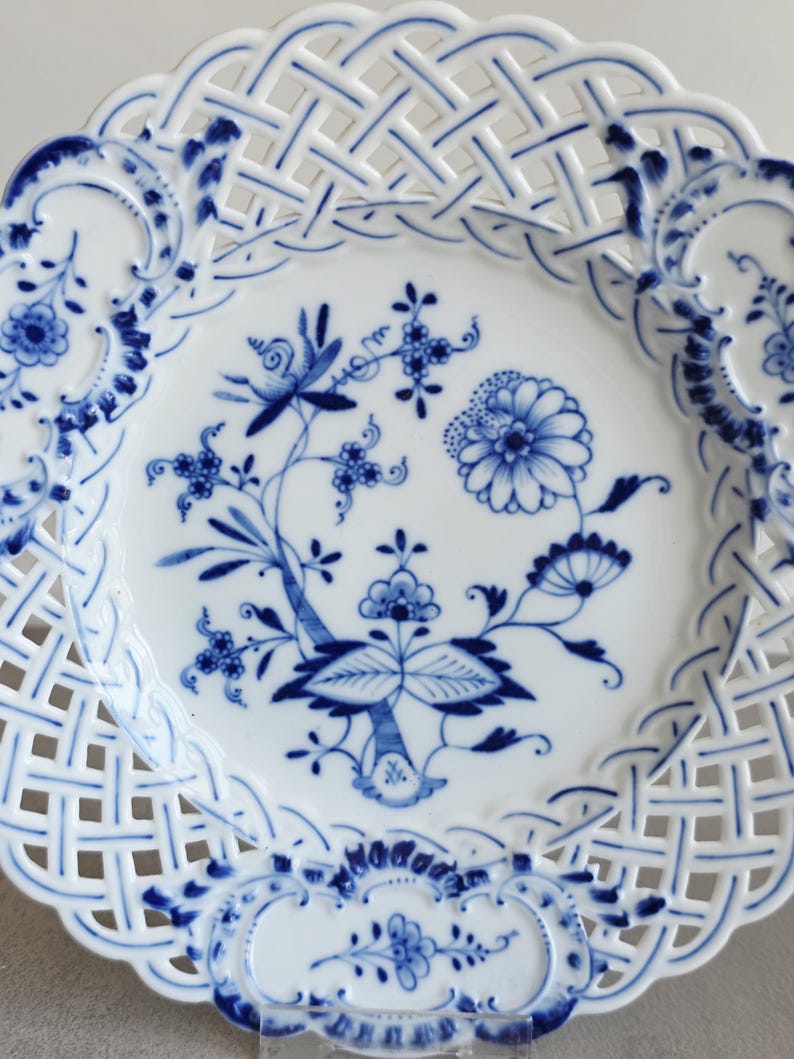 May include: A white porcelain plate featuring a blue floral pattern in the centre and a decorative lattice-style openwork border. The design includes intricate blue painted flowers, leaves, and stems, with scalloped edges and blue accents.