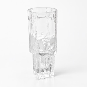 May include: A clear glass vase featuring a cylindrical upper section and a textured, jagged base that resembles melting ice or crystalline formations. The surface of the glass has a rough, organic pattern throughout, creating a light-refracting effect.