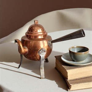 May include: A copper tea kettle with a dark handle and three metal legs. A small ceramic teacup and saucer sit on top of two old books. The kettle has a round body and a lid.