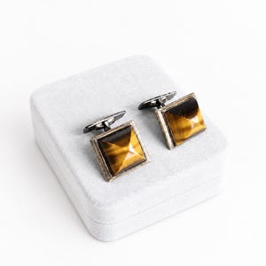 May include: A pair of square cufflinks featuring faceted tiger's eye gemstones set in dark metal frames. Each cufflink has a pyramid-shaped stone with golden-brown chatoyancy and a hinged toggle mechanism on the back for securing to a shirt cuff.