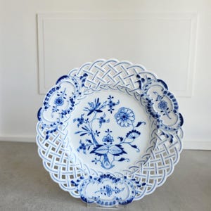 May include: A white porcelain plate featuring a blue floral pattern in the centre and a lattice-style openwork border. The rim is decorated with three distinct blue floral motifs integrated into the woven design of the plate's edge.