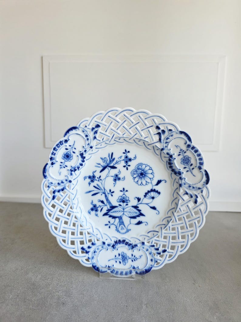 May include: A white porcelain plate featuring a blue floral pattern in the centre and a lattice-style openwork border. The rim is decorated with three distinct blue floral motifs integrated into the woven design of the plate's edge.
