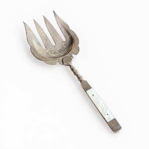 May include: A silver-toned serving fork with a wide, four-tined head featuring engraved scrollwork details. The handle is straight and rectangular, inlaid with a smooth, iridescent white mother-of-pearl panel secured by two small metal rivets.