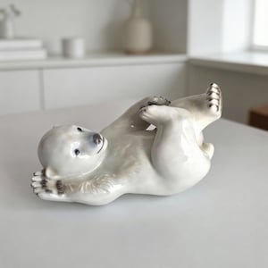 May include: A ceramic polar bear figurine in a playful pose, lying on its back with paws raised. The bear has a white body with grey accents and a light blue nose and eyes. This decorative item is suitable for home decor.