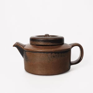 Peut inclure: Une théière en céramique brune avec un corps cylindrique, un bec court et un couvercle assorti. La surface présente un vernis brun terreux et tacheté avec des accents plus sombres, presque noirs, le long du bord du couvercle et de l'épaule.