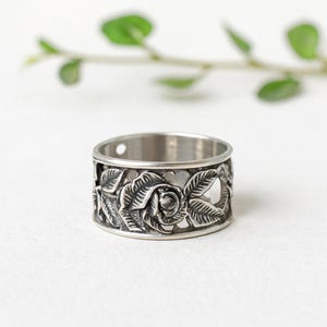 Puede incluir: Un anillo plateado con un diseño floral. El anillo presenta un relieve detallado de una rosa rodeada de hojas. La banda tiene un diseño calado con recortes en forma de corazón. El anillo está sobre un fondo blanco con una rama verde.