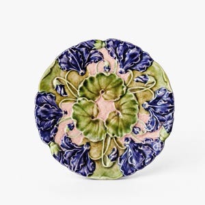 May include: A round ceramic plate featuring a relief design of deep purple morning glory flowers and vibrant green leaves arranged in a circular pattern against a textured, pale pink background with a glossy, crackled glaze finish throughout.