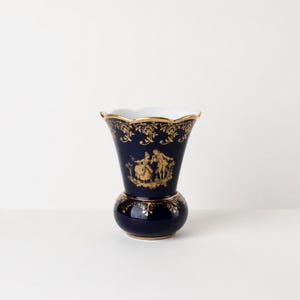 Könnte beinhalten: Eine tief dunkelblaue Porzellanvase mit gewelltem Rand und abgerundetem Boden. Die Außenseite ist mit filigranen, goldfarbenen Blumenmustern und einer zentralen Abbildung zweier Figuren in historischer Kleidung verziert, ergänzt durch Goldränder.