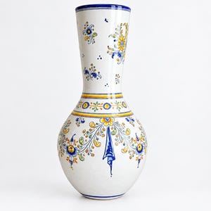 Op de afbeelding: Een hoge keramische vaas met een witte basis en handgeschilderde bloemmotieven in geel, blauw en groen. Het ontwerp bevat gedetailleerde krullende ranken, een blauwe rand en horizontale gele en groene banden rond de hals en het lichaam.