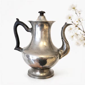 May include: A vintage silver-toned coffee pot with a black handle and spout. The pot has a bulbous body, a lid with a decorative knob, and sits on a pedestal base. The background is white with a sprig of white flowers.