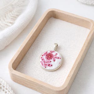 May include: A round pendant with a floral design. The pendant features a white background with a pink floral pattern and a silver-colored metal frame. The pendant is displayed in a light-colored wooden tray.