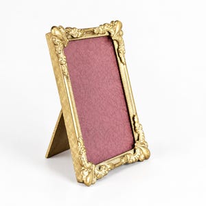 May include: A rectangular standing picture frame with a metallic gold finish. The frame features ornate, scrolled corner detailing and a textured, cross-hatched side panel. It contains a deep burgundy insert with a subtle, repeating pattern.