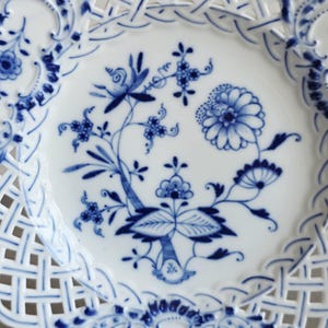 May include: A white porcelain plate featuring a blue floral pattern in the centre and a decorative lattice-style openwork border. The design includes intricate blue painted flowers, leaves, and stems, with a scalloped edge and detailed blue accents.