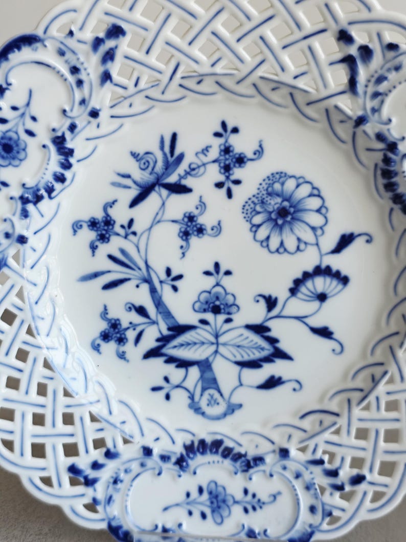 May include: A white porcelain plate featuring a blue floral pattern in the centre and a decorative lattice-style openwork border. The design includes intricate blue painted flowers, leaves, and stems, with a scalloped edge and detailed blue accents.