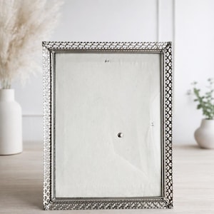 Puede incluir: Un marco de fotos rectangular plateado con un borde decorativo calado. El marco tiene una parte delantera de vidrio y un respaldo blanco. El marco está vacío y listo para exhibir una fotografía o una obra de arte. El marco está sobre una superficie de madera clara.