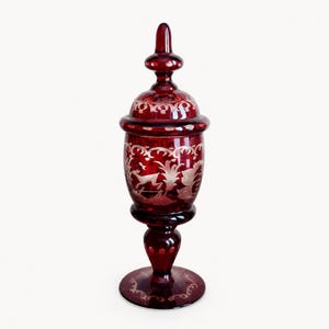 May include: A tall, deep red glass lidded jar featuring intricate etched white designs of a leaping deer and foliage. The vessel has a pedestal base, a rounded body, and a matching removable lid with a pointed finial, all showcasing clear glass.