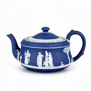 Könnte beinhalten: Eine dunkelblaue Keramik-Teekanne mit weißen Relief-Figuren von Personen und einer Ziege, einem geschwungenen Henkel, einem Ausguss und einem passenden Deckel mit einem weißen Rand in Blattform. Die Kanne hat eine matte Oberfläche.