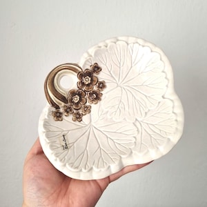 Puede incluir: Una fuente de cerámica blanca con diseño de hojas y un asa marrón adornada con pequeñas flores marrones. El plato se sostiene en una mano sobre un fondo blanco. El texto "Rare Living" es visible.