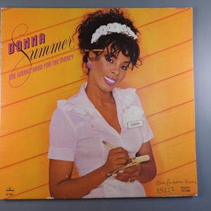 May include: A vinyl record cover featuring Donna Summer in a white waitress uniform with a name tag that reads "Donna". The cover is yellow and orange with the text "Donna Summer She Works Hard for the Money" in pink and white.