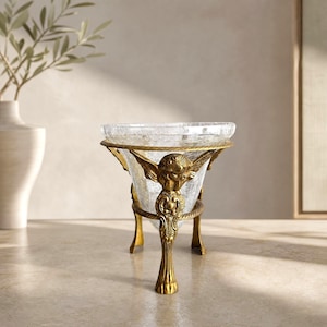 Puede incluir: Un cuenco de cristal craquelado transparente sostenido por un soporte de metal dorado. El soporte tiene tres patas, cada una adornada con una figura de querubín alado esculpido en la parte superior y terminada en un diseño de garra.