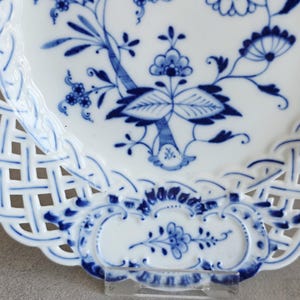 May include: A white porcelain plate featuring a blue floral pattern with intricate botanical motifs. The plate has a decorative lattice-style openwork border with scalloped edges and additional blue floral details along the rim.