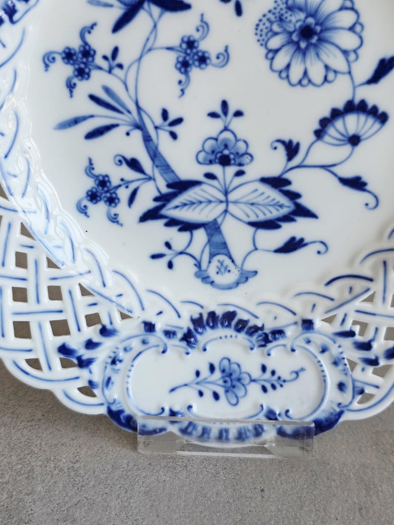 May include: A white porcelain plate featuring a blue floral pattern with intricate botanical motifs. The plate has a decorative lattice-style openwork border with scalloped edges and additional blue floral details along the rim.