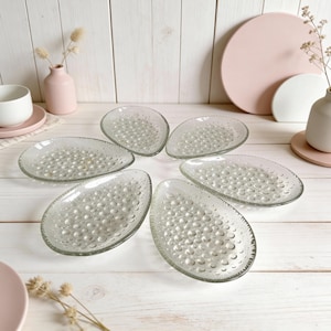 Puede incluir: Seis platos ovalados de vidrio transparente con un patrón texturizado similar a burbujas. Los platos están dispuestos sobre una superficie de madera blanca, con un fondo borroso con elementos decorativos rosas y blancos.