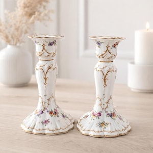 Könnte beinhalten: Zwei weiße Porzellan-Kerzenhalter mit floralen und goldenen Akzenten. Die Kerzenhalter haben einen ausgestellten Sockel und ein detailliertes Design mit bunten Blumen und goldenem Besatz. Das Design umfasst goldene Verzierungen und einen gewellten Rand.