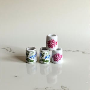 Peut inclure: Cinq petits dés à coudre en céramique blanche. Deux dés à coudre présentent des motifs floraux bleus, tandis que les trois autres affichent des motifs de roses roses. Les dés à coudre sont disposés sur une surface blanche.