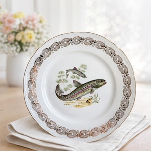 Może przedstawiać: Biały ceramiczny talerz ze zdobionym złotym obramowaniem i szczegółową ilustracją dwóch ryb. Ryby przedstawione są w odcieniach zieleni, fioletu i brązu, z małą rośliną i piaszczystym podłożem. Talerz ma falisty brzeg.