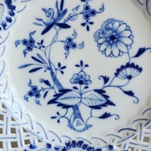 May include: A white ceramic plate featuring a blue floral pattern in the centre and a lattice-style openwork border. The design includes stylised flowers, leaves, and stems painted in shades of blue, with intricate woven details along the edge.