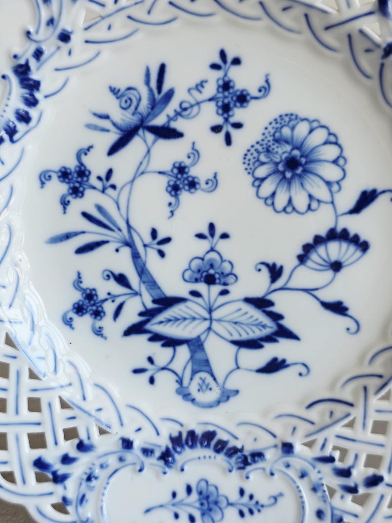 May include: A white ceramic plate featuring a blue floral pattern in the centre and a lattice-style openwork border. The design includes stylised flowers, leaves, and stems painted in shades of blue, with intricate woven details along the edge.