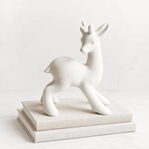 May include: A matte white ceramic figurine of a standing deer with smooth, rounded features and upright ears. The deer is positioned on top of two stacked, light beige hardcover books, showcasing a clean, solid-colored sculptural design.