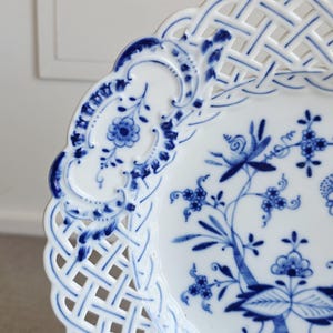 May include: A white porcelain plate featuring a blue floral pattern in the centre and a decorative lattice-style openwork border. The rim is scalloped, and the intricate blue design includes stylised flowers, leaves, and scrolling vines.