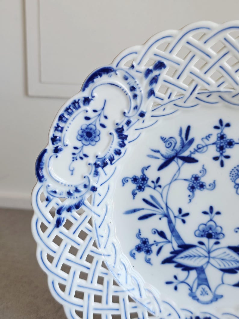 May include: A white porcelain plate featuring a blue floral pattern in the centre and a decorative lattice-style openwork border. The rim is scalloped, and the intricate blue design includes stylised flowers, leaves, and scrolling vines.