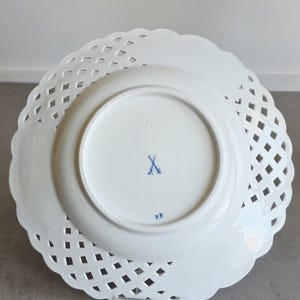 May include: A white porcelain plate with a lattice-style openwork border and a scalloped edge. The underside features a blue crossed-swords maker's mark in the centre and the number 23 printed in blue near the bottom rim of the central base.