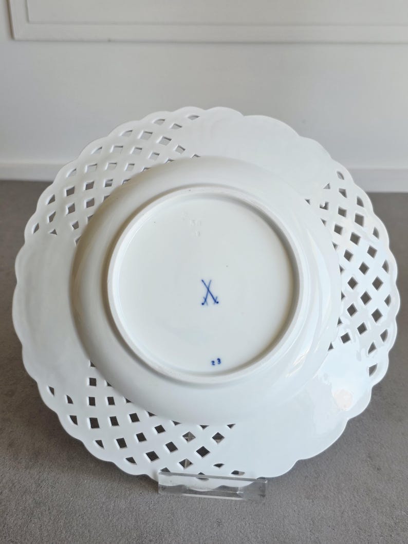 May include: A white porcelain plate with a lattice-style openwork border and a scalloped edge. The underside features a blue crossed-swords maker's mark in the centre and the number 23 printed in blue near the bottom rim of the central base.