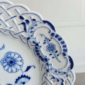 May include: A white porcelain plate featuring a blue floral pattern and a decorative lattice-style openwork border. The rim is scalloped, and the surface is decorated with intricate blue painted flowers and foliage in a classic style.