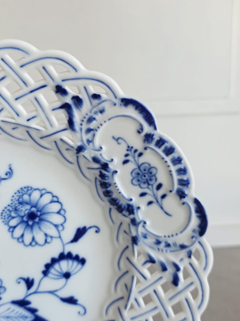 May include: A white porcelain plate featuring a blue floral pattern and a decorative lattice-style openwork border. The rim is scalloped, and the surface is decorated with intricate blue painted flowers and foliage in a classic style.