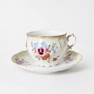 Puede incluir: Un juego de taza de té y platillo de porcelana blanca con un delicado estampado floral de pensamientos en tonos morados, azules y naranjas. El borde de la taza y del platillo tienen una forma festoneada con un acabado en verde suave y dorado.