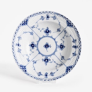 Könnte beinhalten: Ein runder Teller aus weißem Porzellan mit einem tiefblauen Blumenmuster. Das Design umfasst ein zentrales kreisförmiges Motiv mit strahlenden Linien, zarte blaue Blumenzweige und einen gewellten Rand mit schuppenartigen blauen Details.