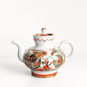 May include: A white porcelain teapot featuring a hand-painted design of a rooster and floral motifs in shades of orange, green, and gold. The teapot has a rounded body, a curved spout, a looped handle, and a lid with a small gold-trimmed knob.