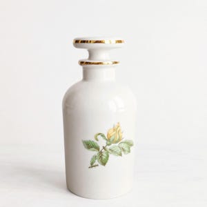May include: A white porcelain apothecary jar with a cylindrical body and a matching stopper. The jar features a hand-painted yellow rosebud with green leaves on the front and gold-painted trim on the rim of the stopper and the jar neck.